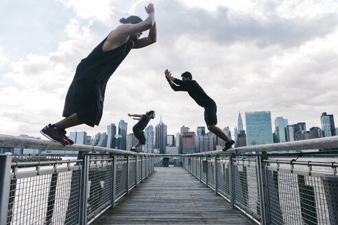 USA, New York, Brooklyn, drei junge Männer machen Parkour-Sprünge auf dem Pier vor der Skyline von Manhattan - JUBF00336