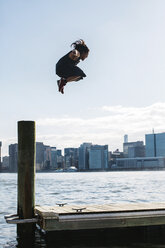 USA, New York, Brooklyn, young man doing Parkour jump from wooden pole in front of Manhattan skyline - JUBF00340