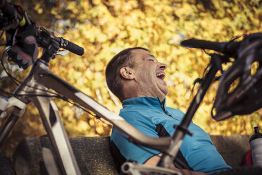 Lachender Mann mit Mountainbike, der auf einer Bank sitzend eine Pause macht - SEBF00060