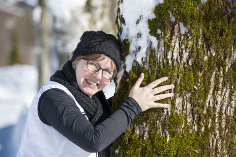 Porträt einer lächelnden Frau, die sich im Winter an einen Baum schmiegt, lizenzfreies Stockfoto