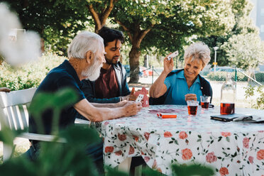 Cheerful senior woman playing cards with males at table in back yard - MASF11526