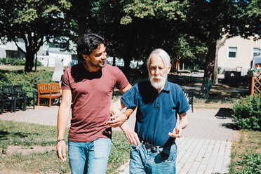 Lächelnder männlicher Pfleger, der Arm in Arm mit einem älteren Mann im Pflegeheim spazieren geht - MASF11532