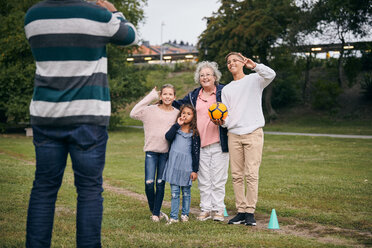 Mittelteil eines Mannes, der eine glückliche Großmutter und Enkelkinder fotografiert, die während eines Picknicks im Park ein Friedenszeichen machen - MASF11536