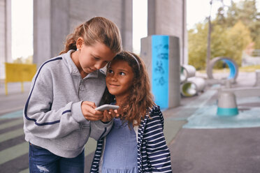 Lächelnde Schwestern, die ein Mobiltelefon benutzen, während sie auf dem Fußweg eines Spielplatzes stehen - MASF11539