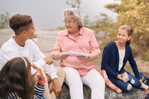 Happy grandmother giving gift to grandson while sitting with granddaughters in park during picnic stock photo