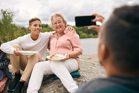 Man photographing grandson and grandmother with meal on rock during picnic stock photo