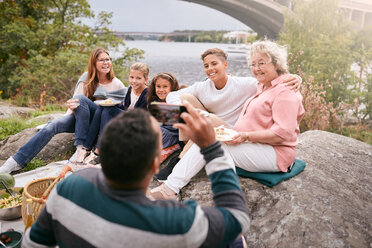 Mann fotografiert glückliche Familie mit Essen auf einem Felsen im Park während eines Picknicks - MASF11552