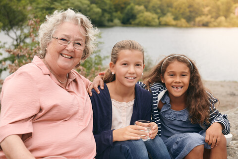 Porträt von lächelnden Enkeltöchtern und Großmutter am Seeufer im Park, lizenzfreies Stockfoto