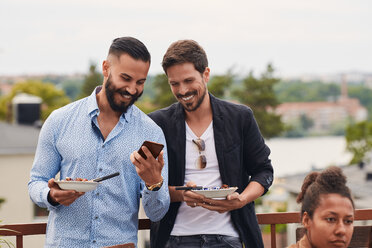 Ein lächelnder Mann zeigt einem Freund sein Handy, während er auf einer Terrasse eine Mahlzeit einnimmt - MASF11572