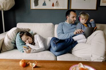 Mädchen, das ein Mobiltelefon benutzt, während der Vater seiner Schwester auf der Couch im Wohnzimmer ein digitales Tablet zeigt - MASF11587