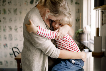 Liebevolle Tochter, die ihre Mutter umarmt, während sie zu Hause auf dem Küchentisch sitzt - MASF11593