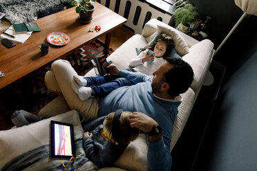 Hochformatige Ansicht von Vater und Töchtern, die verschiedene Technologien auf der Couch im Wohnzimmer zu Hause verwenden - MASF11617