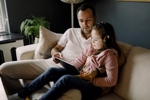 Vater hilft seiner Tochter bei der Benutzung eines digitalen Tablets auf der Couch zu Hause, lizenzfreies Stockfoto