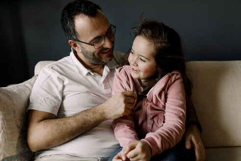 Lächelnder Vater sieht seine Tochter an, während er auf der Couch im Wohnzimmer sitzt - MASF11626