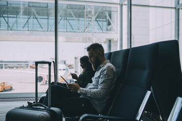 Colleagues using technologies while sitting in waiting room at airport departure area - MASF11656