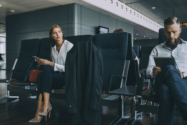 Nachdenkliche junge Geschäftsfrau, die wegschaut, während sie mit einem Kollegen im Abflugbereich des Flughafens sitzt - MASF11658