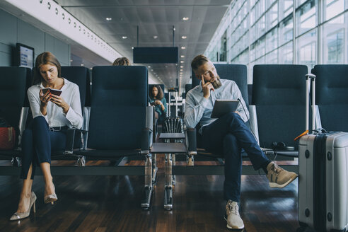 Nachdenklicher Geschäftsmann schaut weg, während er neben einer Kollegin im Wartebereich eines Flughafens sitzt - MASF11660
