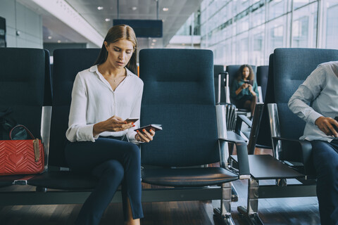 Junge Geschäftsfrau, die mit ihrem Smartphone Textnachrichten verschickt, während sie mit ihrem Reisepass im Wartezimmer des Flughafens sitzt, lizenzfreies Stockfoto