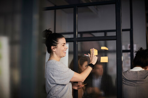Lächelnde kreative Geschäftsfrau klebt Haftnotiz auf Glas im Büro - MASF11694