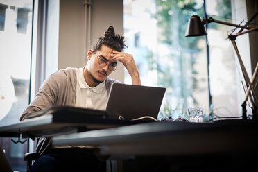 Angespannter Geschäftsmann, der auf seinen Laptop schaut, während er in einem kreativen Büro sitzt - MASF11718