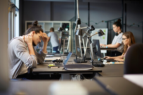 Angespannter männlicher Unternehmer, der auf seinen Laptop schaut, während er in einem kreativen Büro sitzt, lizenzfreies Stockfoto