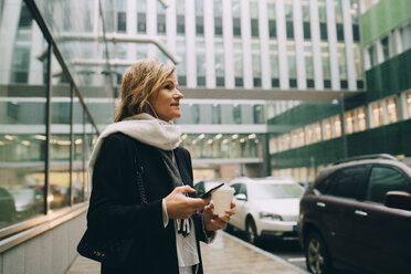 Geschäftsfrau mit Kaffee und Smartphone schaut weg, während sie auf dem Bürgersteig vor einem modernen Gebäude in der Stadt steht - MASF11748