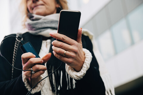 Mittelteil einer Geschäftsfrau, die ein Mobiltelefon mit Ladegerät hält, während sie an einem Gebäude in der Stadt steht, lizenzfreies Stockfoto