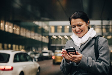 Lächelnde Geschäftsfrau, die ein Smartphone benutzt, während sie auf der Straße vor einem Gebäude steht - MASF11751