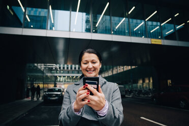 Lächelnde Geschäftsfrau, die ein Mobiltelefon benutzt, während sie auf der Straße vor einem Gebäude in der Stadt steht - MASF11756