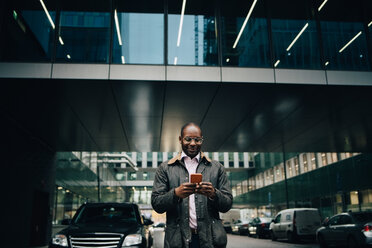 Low angle view of businessman using smart phone while standing on road against building in city - MASF11759