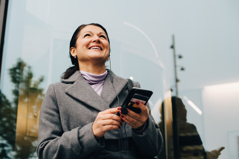 Niedriger Winkel Blick auf lächelnde Geschäftsfrau mit Handy, während stehend gegen Gebäude in der Stadt, lizenzfreies Stockfoto