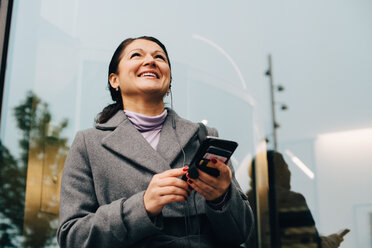 Low angle view of smiling businesswoman using mobile phone while standing against building in city - MASF11765