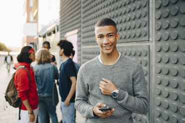 Porträt eines lächelnden Teenagers, der ein Handy hält, während seine Freunde im Hintergrund auf der Straße stehen - MASF11778