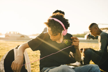 Jugendlicher hört Musik über Kopfhörer, während er mit einem Freund an einem sonnigen Tag sitzt - MASF11781