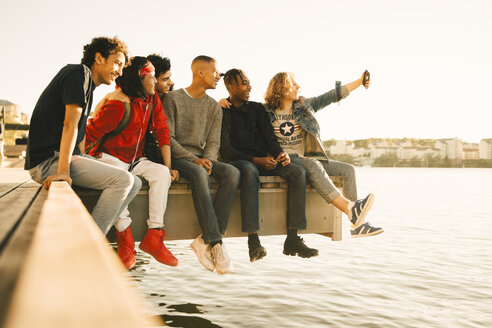 Glückliche männliche Freunde, die ein Selfie machen, während sie Seite an Seite auf dem Pier am Fluss sitzen - MASF11785