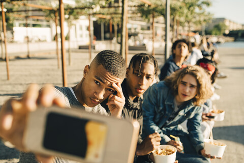 Männliche Freunde, die ein Selfie machen, während sie auf der Straße in der Stadt Essen zum Mitnehmen essen, lizenzfreies Stockfoto