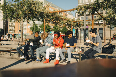 Männliche Freunde essen Essen zum Mitnehmen und sitzen nebeneinander auf einer Bank in der Stadt - MASF11797
