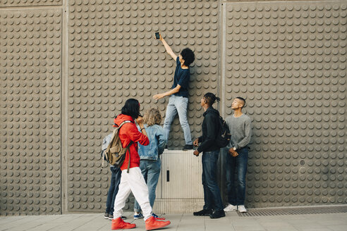 In voller Länge von männlichen Freunden, die ein Selfie mit einem Mobiltelefon machen, während sie an einer Mauer in der Stadt stehen - MASF11811