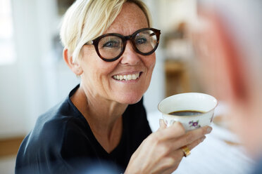 Lächelnde reife Frau, die eine Kaffeetasse hält, während sie zu Hause wegschaut - MASF11829