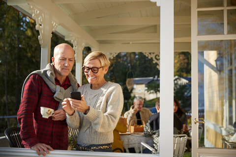 Cheerful mature woman showing mobile phone to bald man with friends in background on porch stock photo