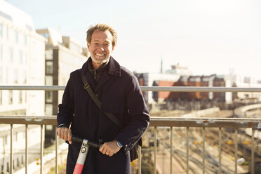 Portrait of smiling mature man standing with electric push scooter on bridge against sky in city - MASF11863