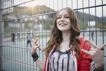 Porträt eines glücklichen Teenagers, der an einem Zaun auf einem Sportplatz gestikuliert - RORF01826