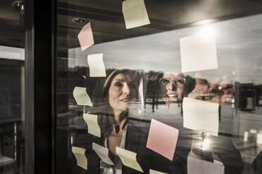 Two businesswomen brainstorming, putting sticky notes on window pane - MJRF00083