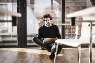 Businessman sitting on floor of his office, using laptop and headphones - MJRF00115