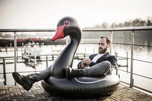 Hipper Geschäftsmann arbeitet auf einem Hausboot, sitzt auf einem schwimmenden Schwan und benutzt eine Smartwatch - MJRF00133