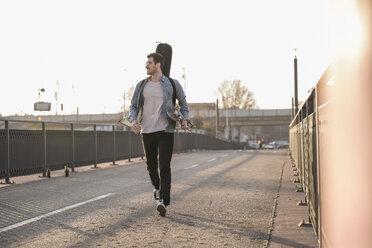 Smiling young man with guitar case and skateboard walking on a bridge in the city - UUF16790