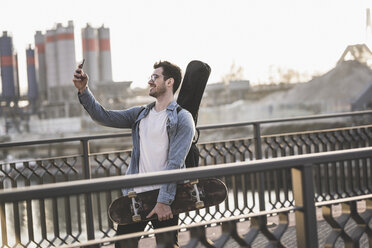 Junger Mann mit Gitarrenkoffer und Skateboard macht ein Selfie in der Stadt - UUF16792