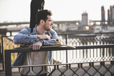 Junger Mann mit Gitarrenkoffer und Skateboard auf einer Brücke in der Stadt und schaut sich um - UUF16793