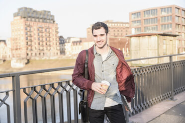 Lächelnder junger Mann mit Tablet, Tasche und Kaffee zum Mitnehmen auf einer Brücke in der Stadt - UUF16794