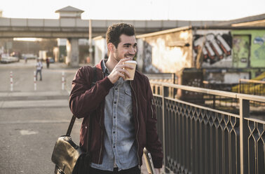 Lächelnder junger Mann mit Tablet, Tasche und Kaffee zum Mitnehmen auf einer Brücke in der Stadt - UUF16798